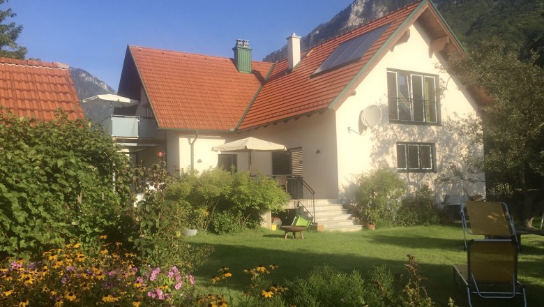 Ein Haus mit rotem Dach, umgeben von einem Garten mit Blumen und Liegest&uuml;hlen, vor einer Bergkulisse.