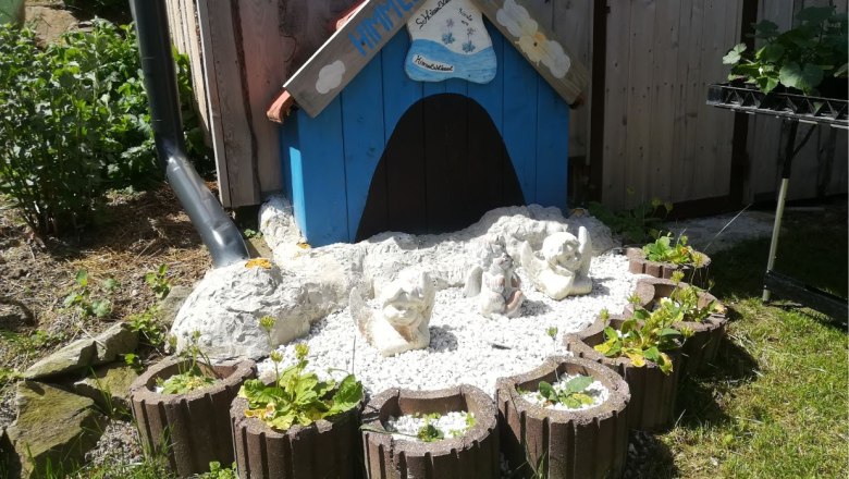 Kleines blaues Häuschen mit Engelfiguren und Pflanzen in einem Garten.