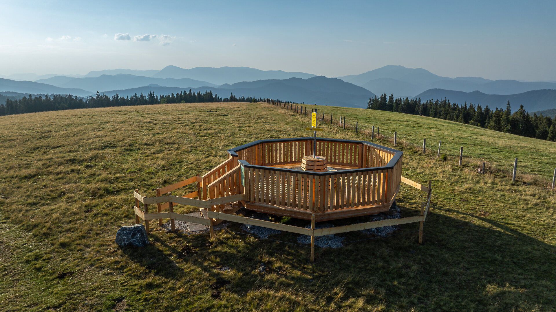 Hölzerner Aussichtsplatz auf einer weitläufigen Almwiese mit Blick auf die umliegende Berglandschaft.