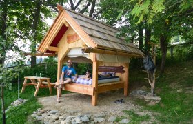 Ein kleines Holzhaus mit einem Bett im Freien, umgeben von Bäumen. Zwei Personen sitzen und liegen auf dem Bett. Ein Picknicktisch steht daneben.