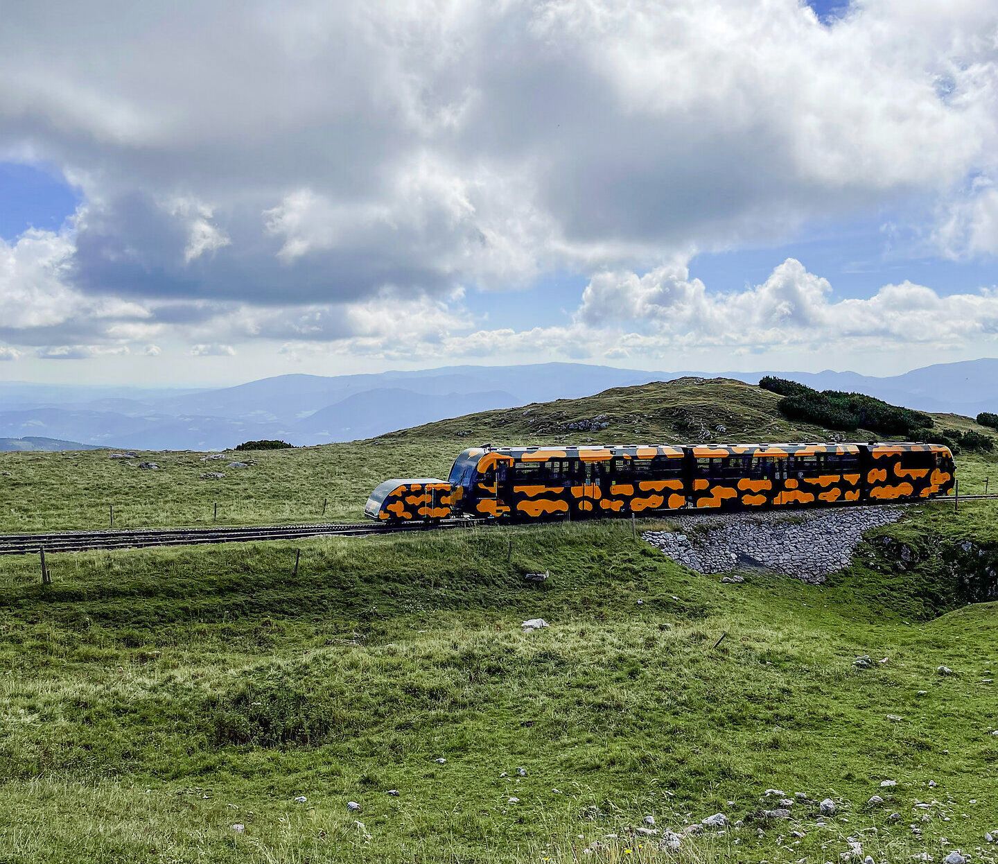 Ein farbenfroher Zug schlängelt sich durch die sanften Hügel der Wiener Alpen, umgeben von üppigem Grün und majestätischen Ausblicken. Die Wolken ziehen gemächlich über den Himmel und verleihen der Szenerie eine friedliche Atmosphäre, die zum Verweilen einlädt.