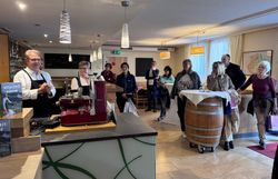 Rund 8 Personen stehen in einer Gaststube und hören den Gastgebern, die hinter der Bar etwas erzählen, zu. 