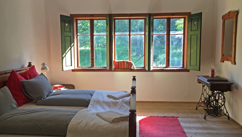 Cozy double room with wooden floor, bed, window with green shutters and a red carpet.