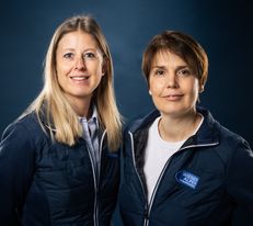 Portraitfoto von zwei Wiener Alpen Mitarbeiterinnen, die eine Jacke mit Wiener Alpen Logo tragen und vor einem dunklem Hintergrund stehen.