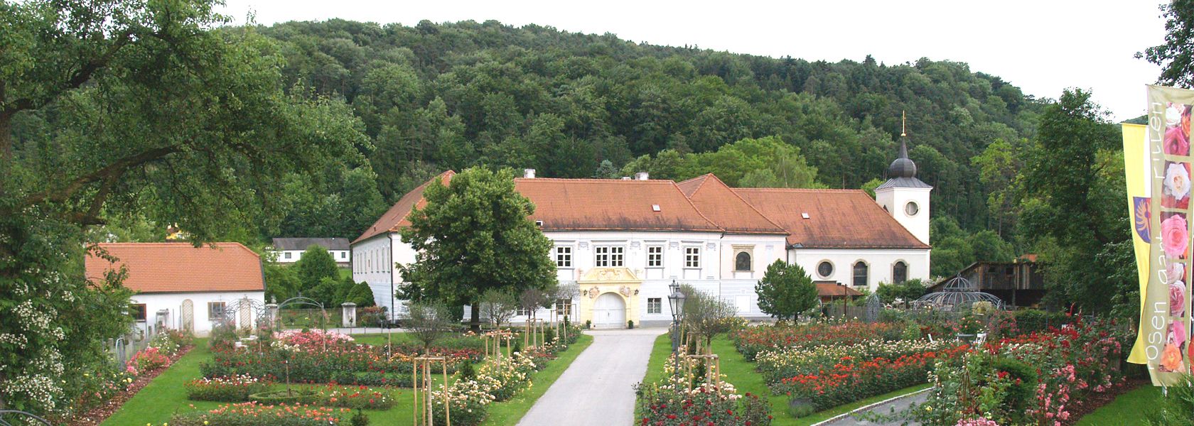 Ein historisches Gebäude mit rotem Dach, umgeben von einem Rosengarten und Bäumen, im Hintergrund ein bewaldeter Hügel.