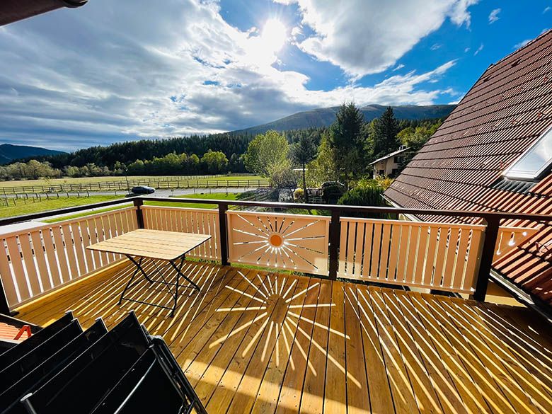 Wooden balcony with table, view of meadows and mountains, sunbeam pattern on the floor.
