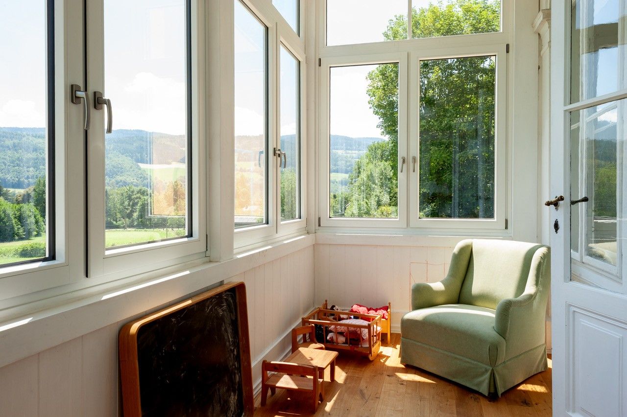 Eine helle Veranda mit großen Fenstern, einem grünen Sessel und Kinderspielzeug auf Holzboden.