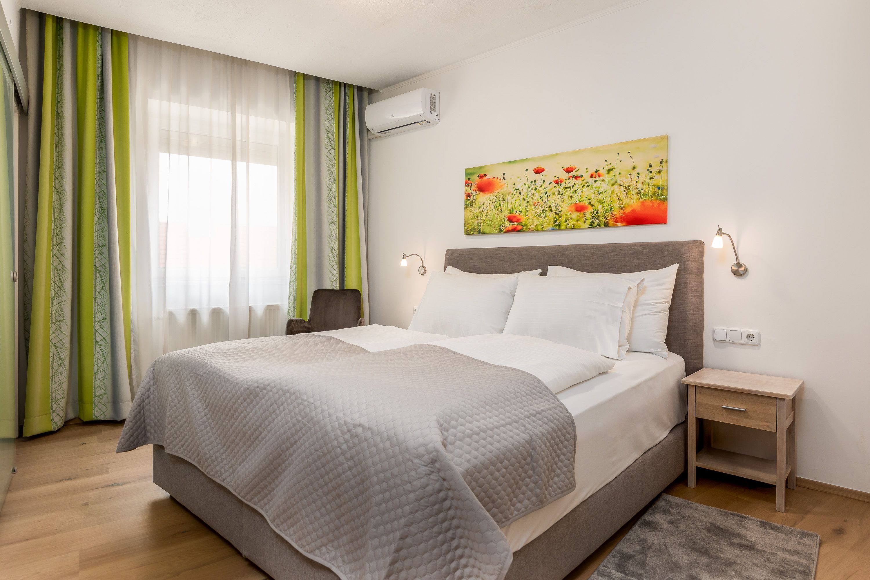 Helles Zimmer mit Doppelbett, grünen Vorhängen, Teppich, Holzboden, Nachtkästchen, Klimaanlage und Sitzmöglichkeiten