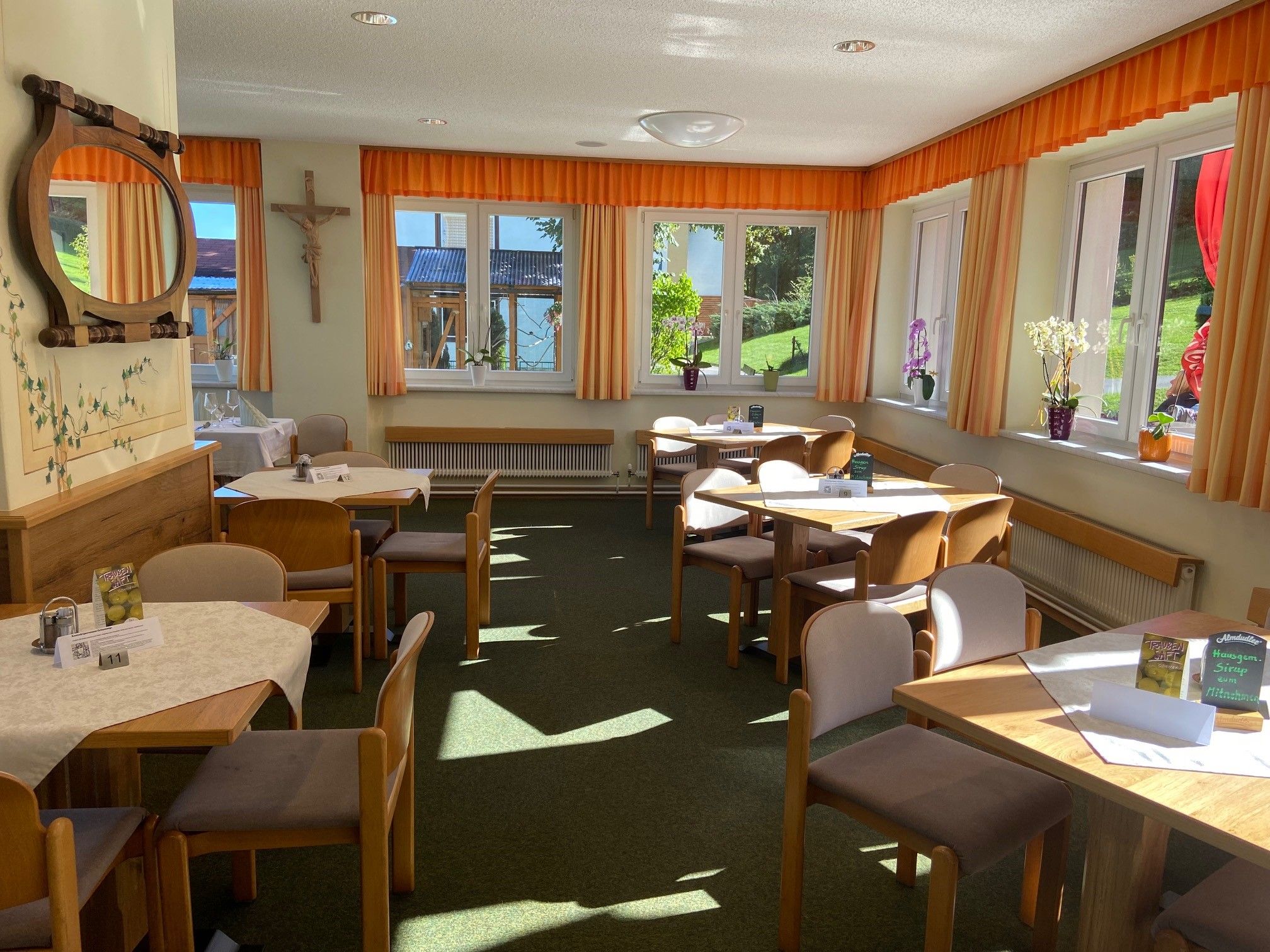 Ein heller Speisesaal mit Holztischen, Stühlen und großen Fenstern mit orangefarbenen Vorhängen.