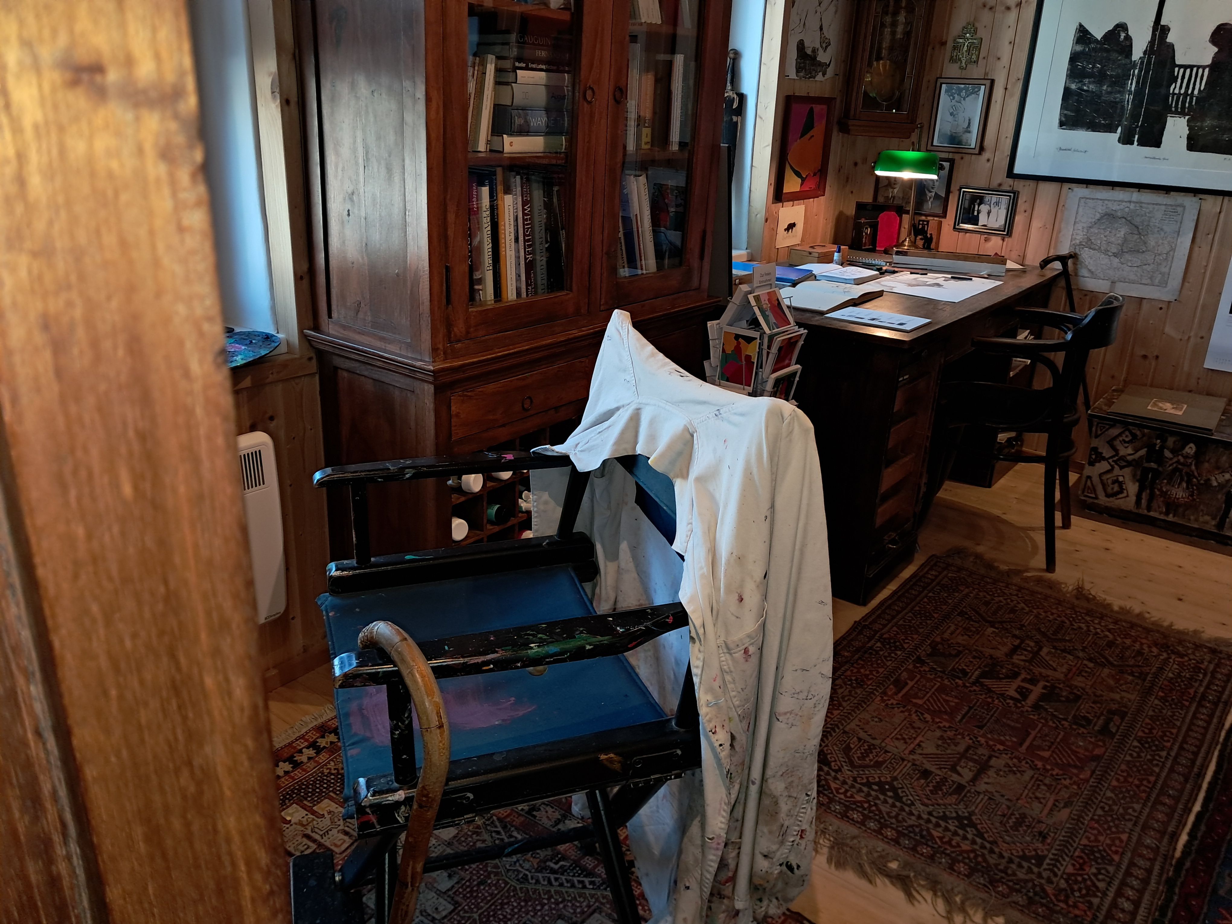 Ein Künstleratelier mit einem bemalten Stuhl, einem Schreibtisch voller Papiere und einem Bücherregal im Hintergrund.