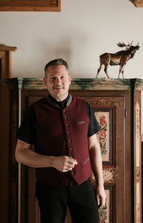 Mann in weinroter Trachten-Weste steht in einer gemütlichen Stube vor einem kunstvoll verzierten Holzschrank.