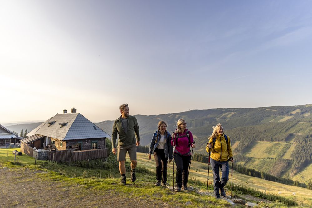 Vier Personen wandern auf einem Bergweg neben einer Hütte bei Sonnenuntergang.