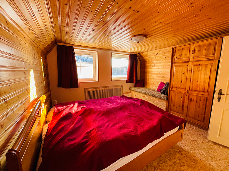Wood-paneled bedroom with red comforter cover, two windows, a wardrobe and a bench.