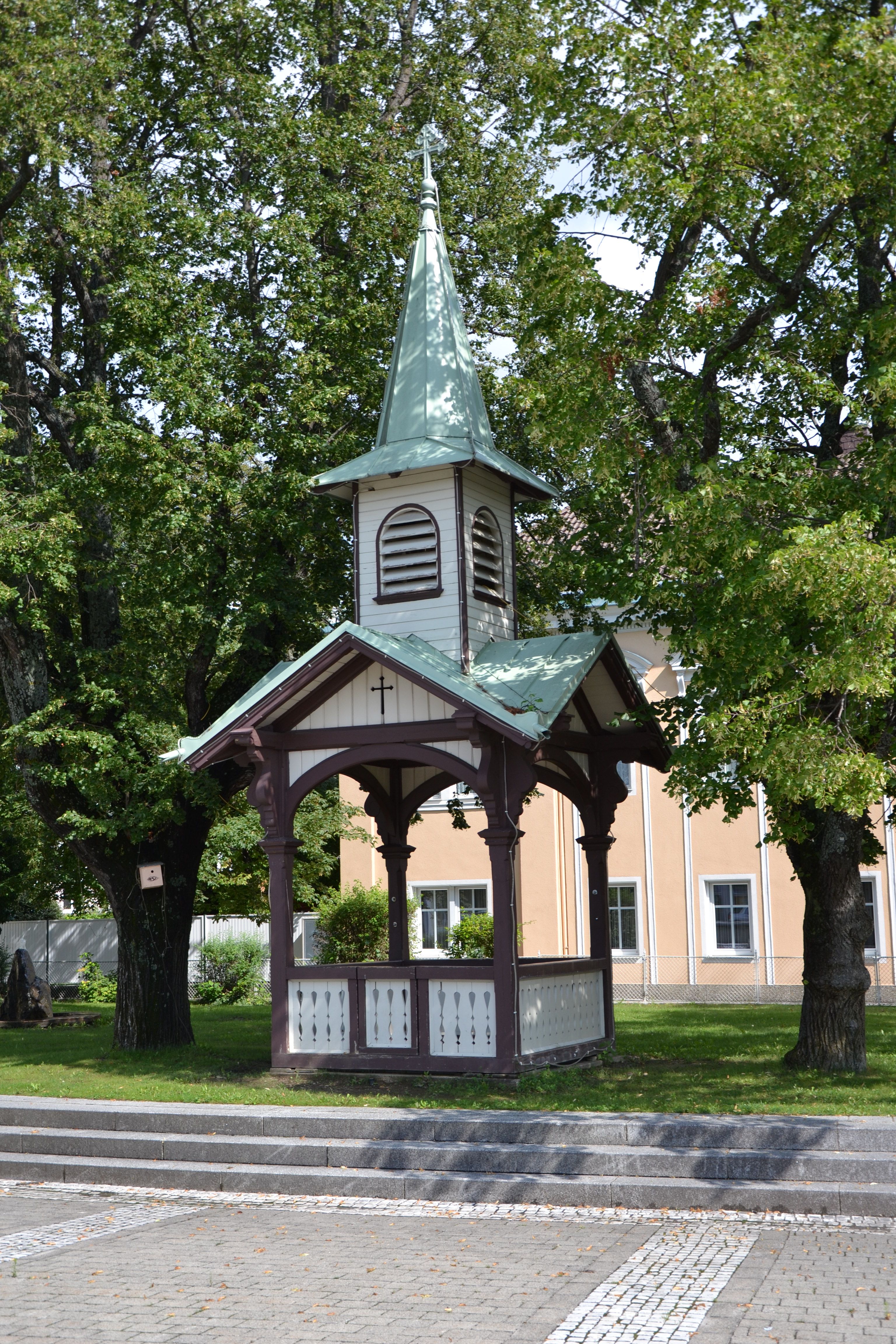 Ein kleiner Pavillon mit spitzem Dach und Kreuz, umgeben von Bäumen.