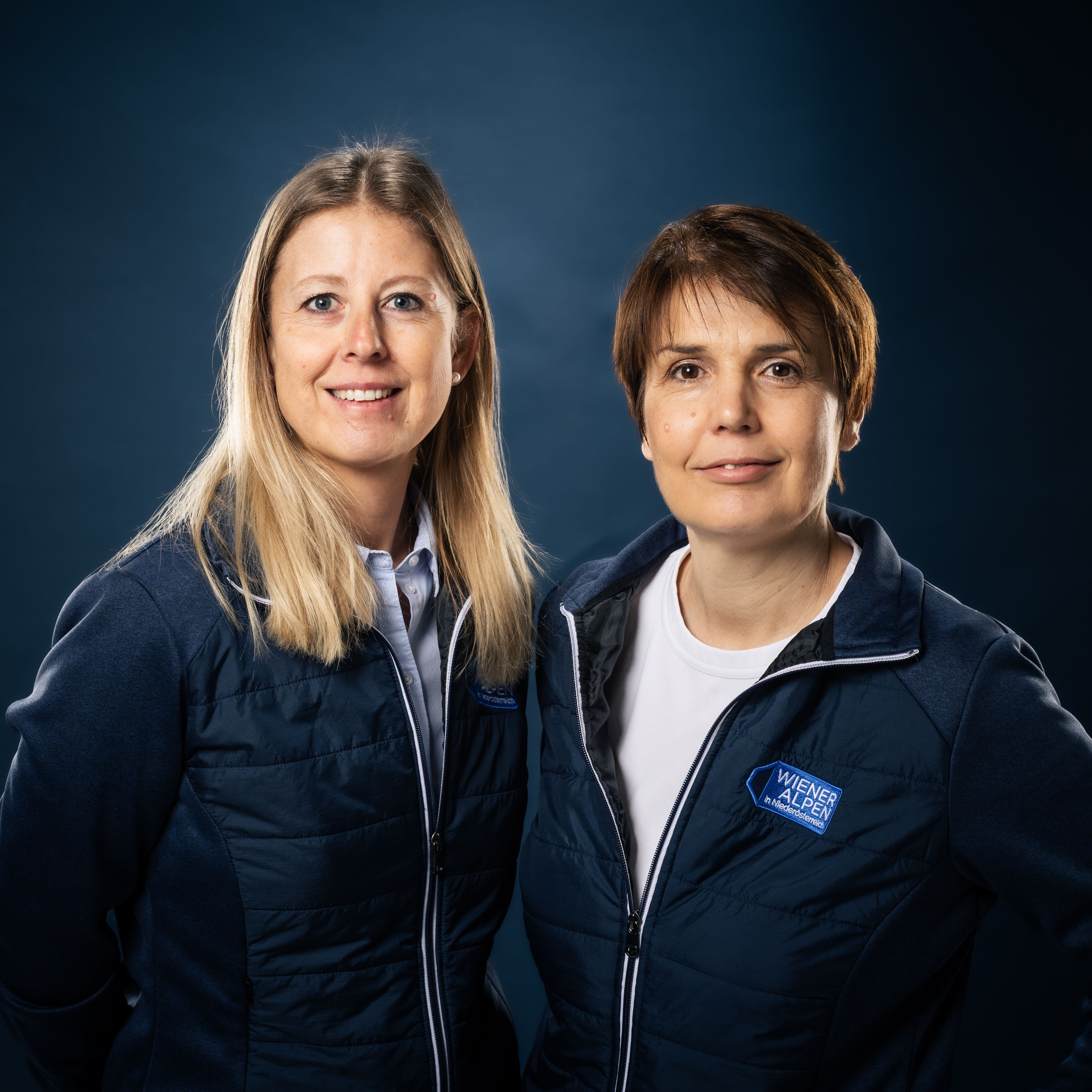 Portraitfoto von zwei Wiener Alpen Mitarbeiterinnen, die eine Jacke mit Wiener Alpen Logo tragen und vor einem dunklem Hintergrund stehen.