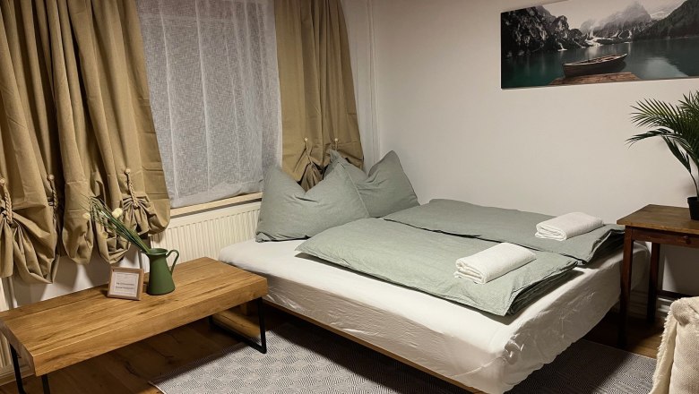 Gemütliches Schlafzimmer mit Doppelbett, grüner Bettwäsche, Holzmöbeln und Pflanzen. Ein Bild von einem See hängt an der Wand.