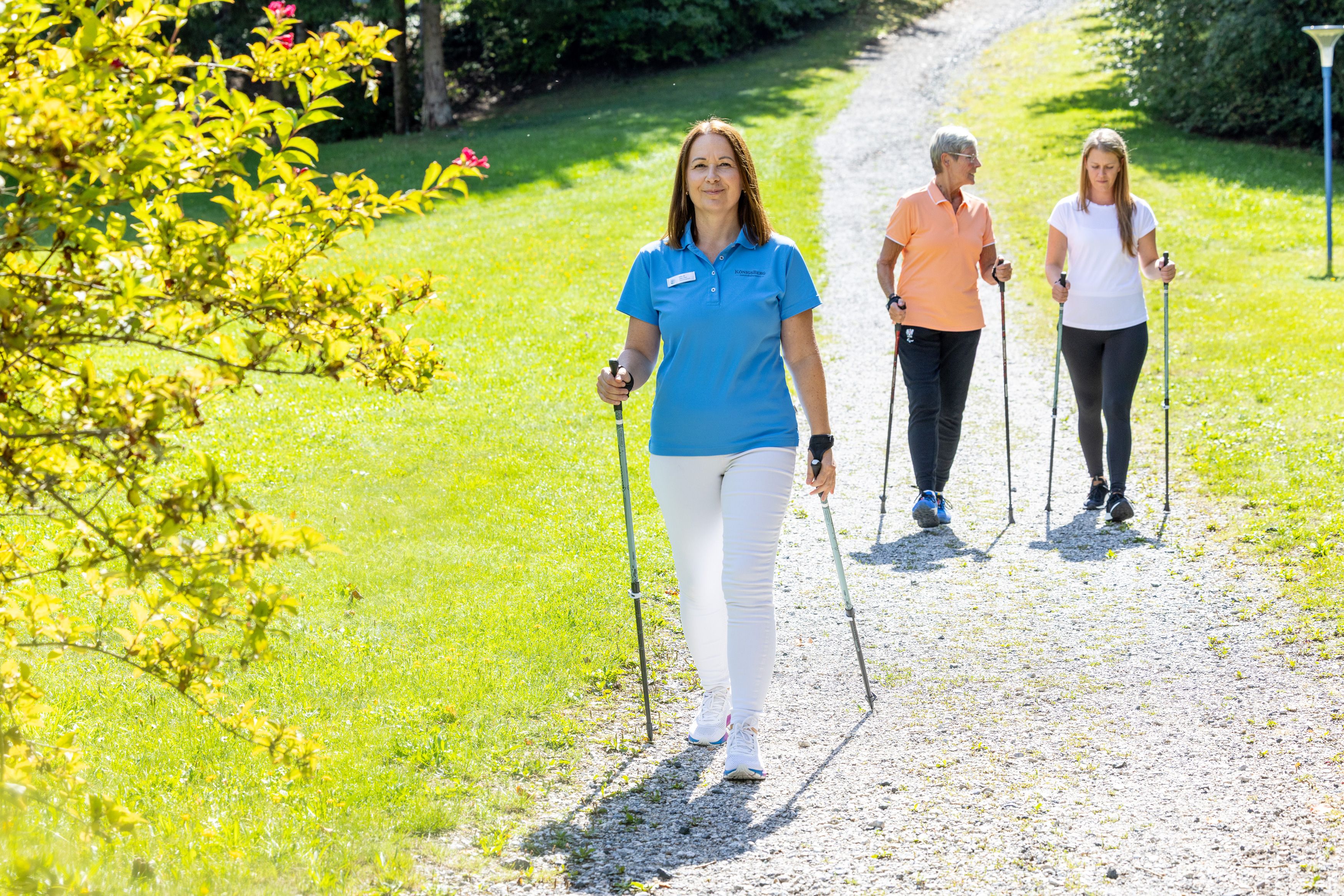 Drei Frauen beim Nordic Walking auf einem sonnigen Weg im Grünen.