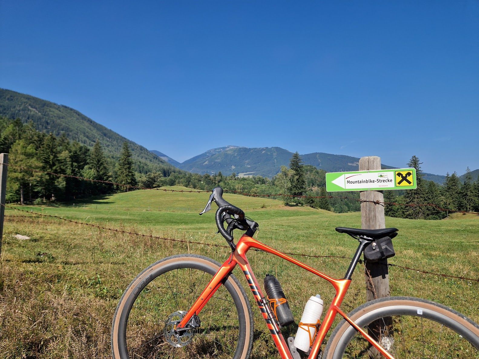 Ein orangefarbenes Fahrrad steht vor einer grünen Wiese mit einem Schild, das auf eine Mountainbike-Strecke hinweist. Im Hintergrund sind bewaldete Berge zu sehen.