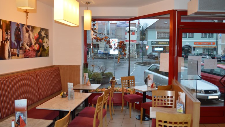 Innenansicht eines gemütlichen Cafés mit roten Stühlen und Tischen, Blick auf eine Straße durch große Fenster.