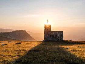 Die sanften H&uuml;gel der Wiener Alpen umarmen die kleine, charmante Kapelle, die im goldenen Licht der untergehenden Sonne erstrahlt. Ihre silhouette hebt sich majest&auml;tisch gegen den Himmel ab und l&auml;dt Wanderer ein, einen Moment der Stille und Besinnung zu genie&szlig;en. Hier, wo die Natur und die Spiritualit&auml;t harmonisch verschmelzen, wird jeder Besuch zu einem unvergesslichen Erlebnis.