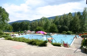 Freibad mit Schwimmern und Sonnenschirmen, umgeben von Bäumen und Hügeln.