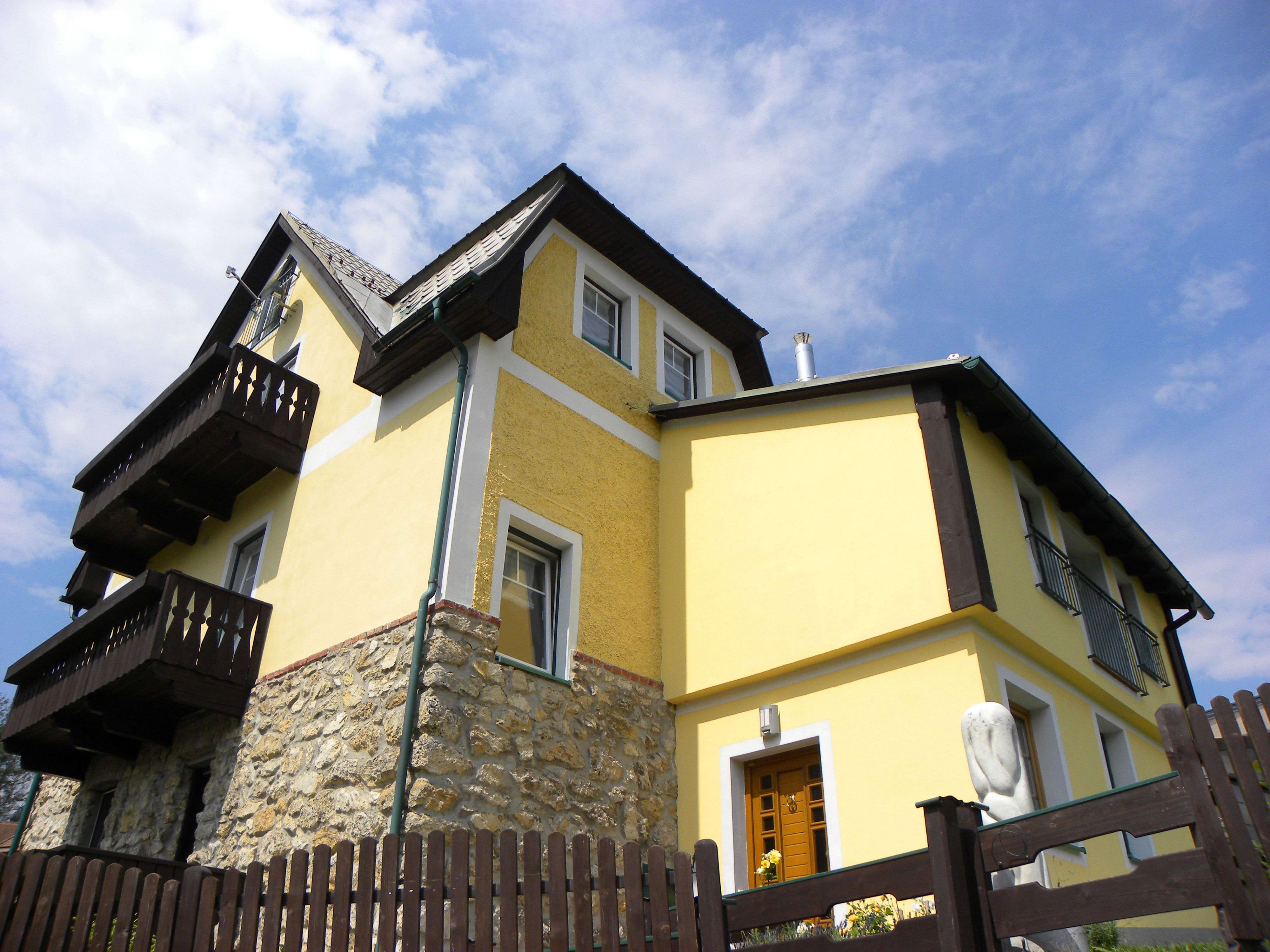 Yellow house with wooden details and stone wall, blue sky in the background.