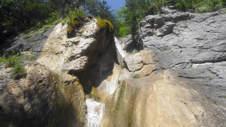 Wasserfall in felsiger Landschaft mit Bäumen und blauem Himmel.