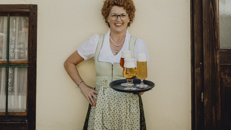 Frau in traditioneller Kleidung mit Tablett voller Biergl&auml;ser vor einer Wand.