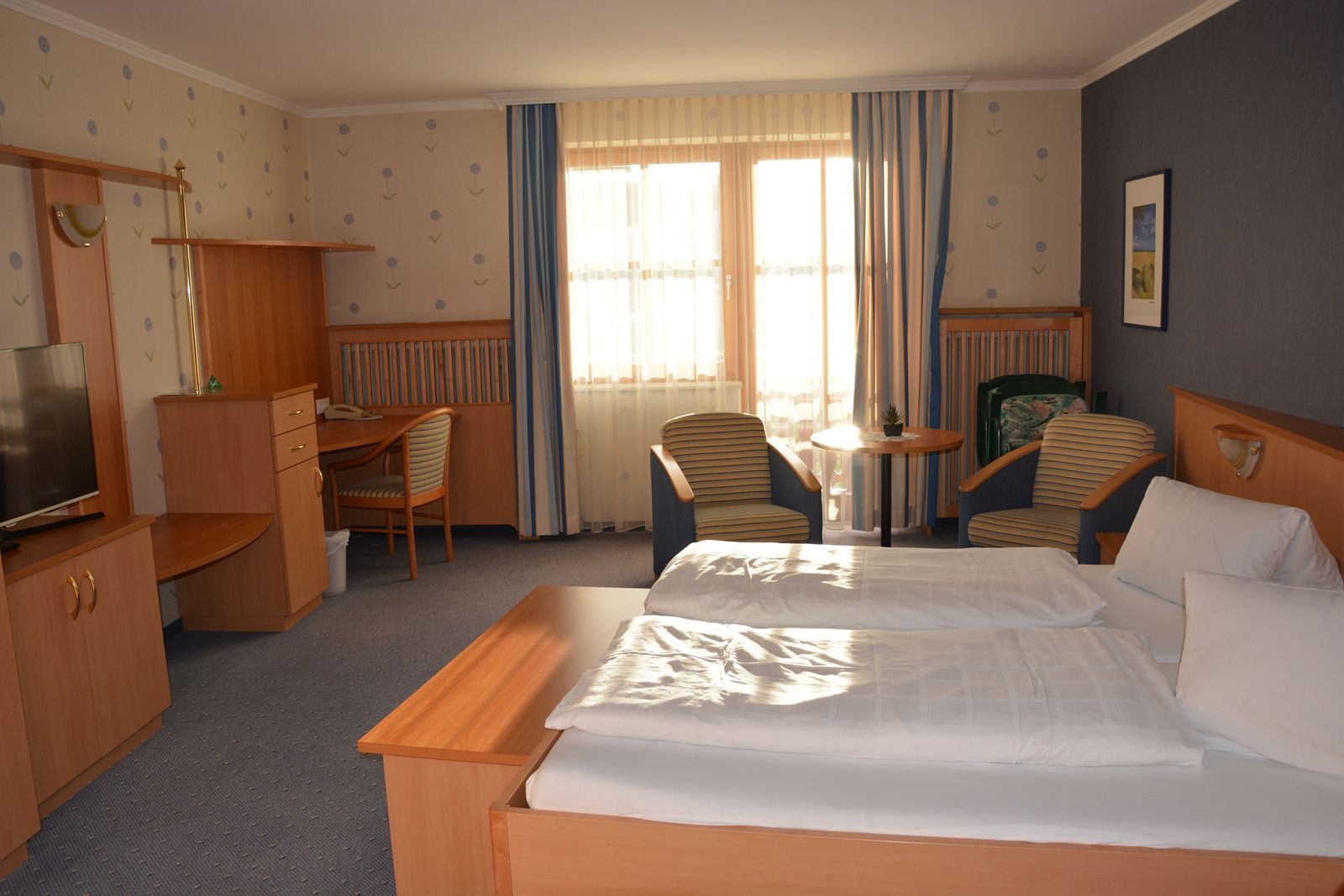 Ein gemütliches Hotelzimmer mit Doppelbett, Schreibtisch, Fernseher und zwei Sesseln vor einem Fenster mit Vorhängen.