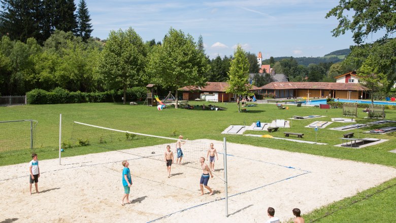 Kinder spielen Volleyball auf einem Sandplatz im Freibad Aspang, umgeben von grüner Wiese und Bäumen.