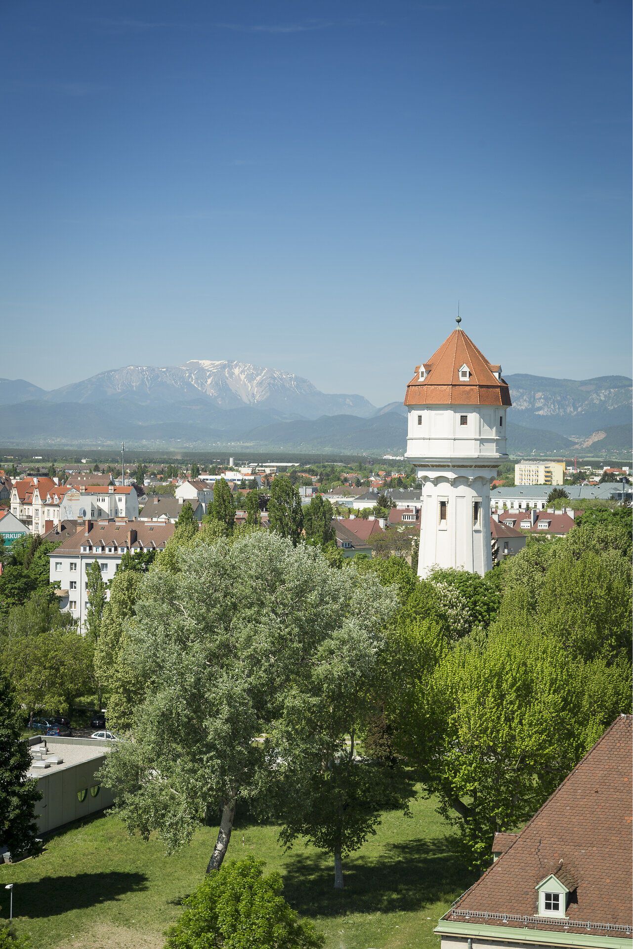 Der historische Wasserturm von Wiener Neustadt erhebt sich über den Baumwipfeln. Im Hintergrund sind die Dächer der Stadt und das Schneebergmassiv zu sehen.