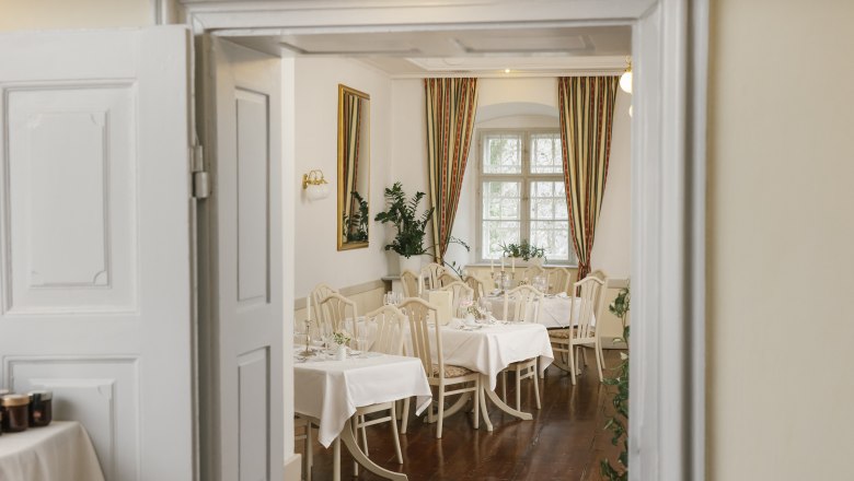 Elegantes Restaurant mit gedeckten Tischen, weißen Tischdecken und klassischen Stühlen, durch eine geöffnete Tür sichtbar.