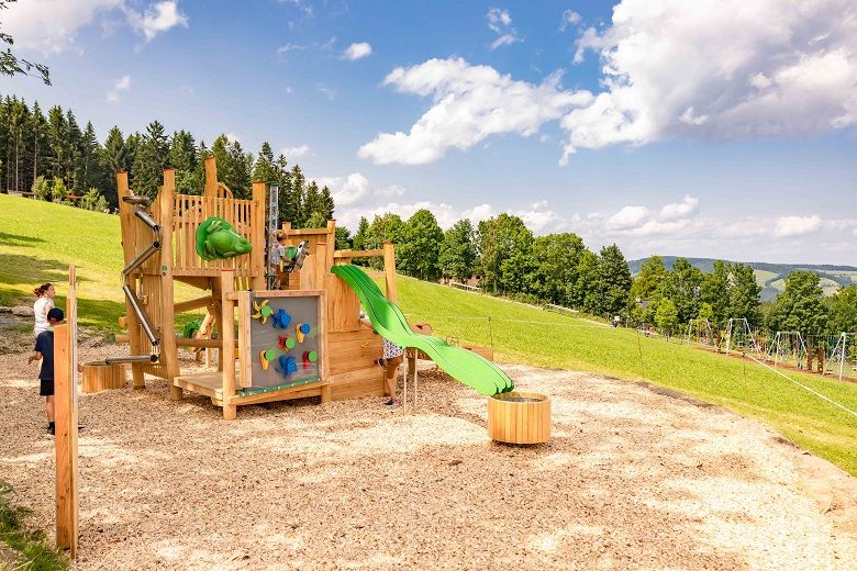 Spielplatz mit Klettergerüst und Rutsche auf einer Wiese, umgeben von Bäumen und blauem Himmel.