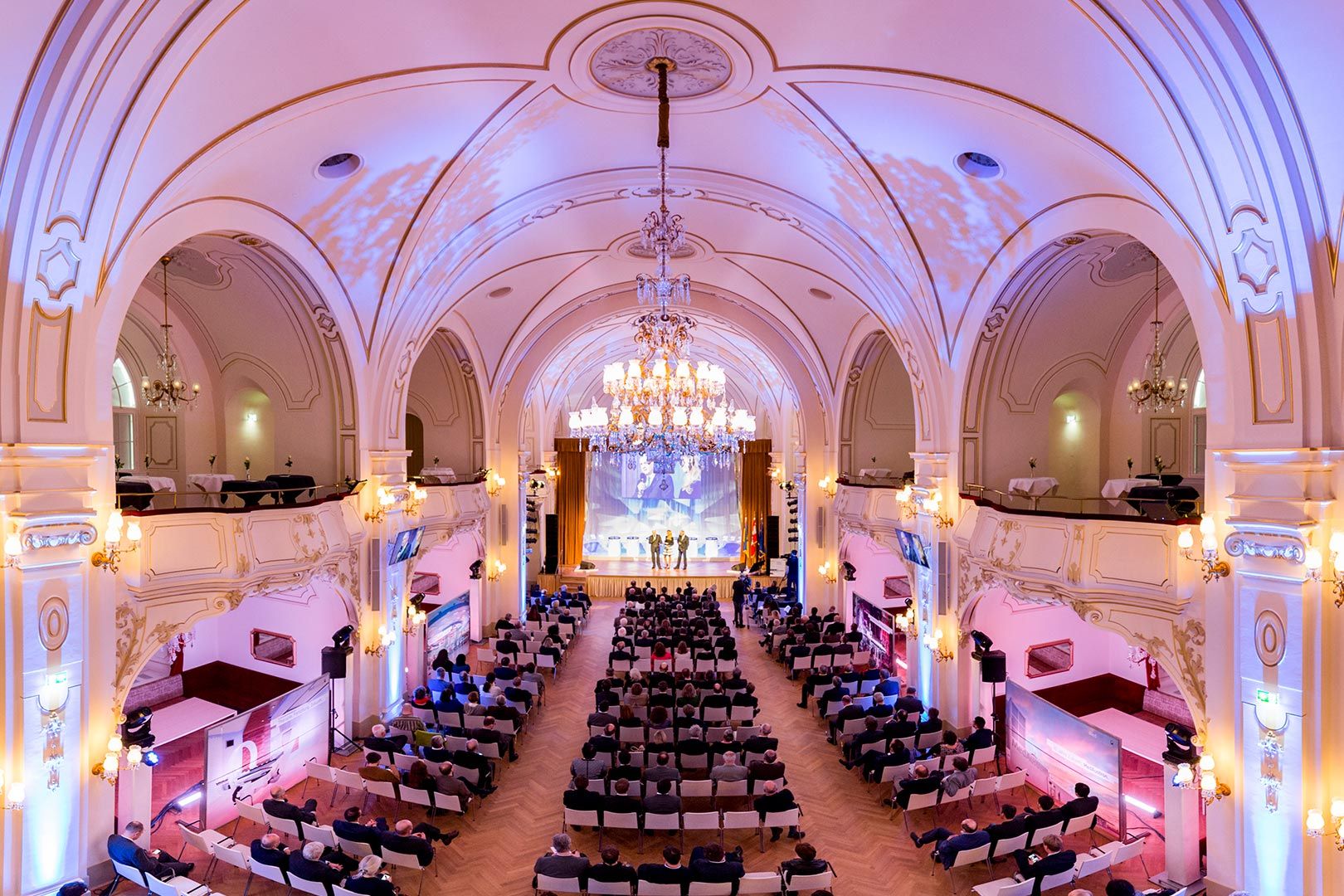 Prunkvoller, weiß-goldener Saal mit Bühne, Bestuhlung und großer Luster.