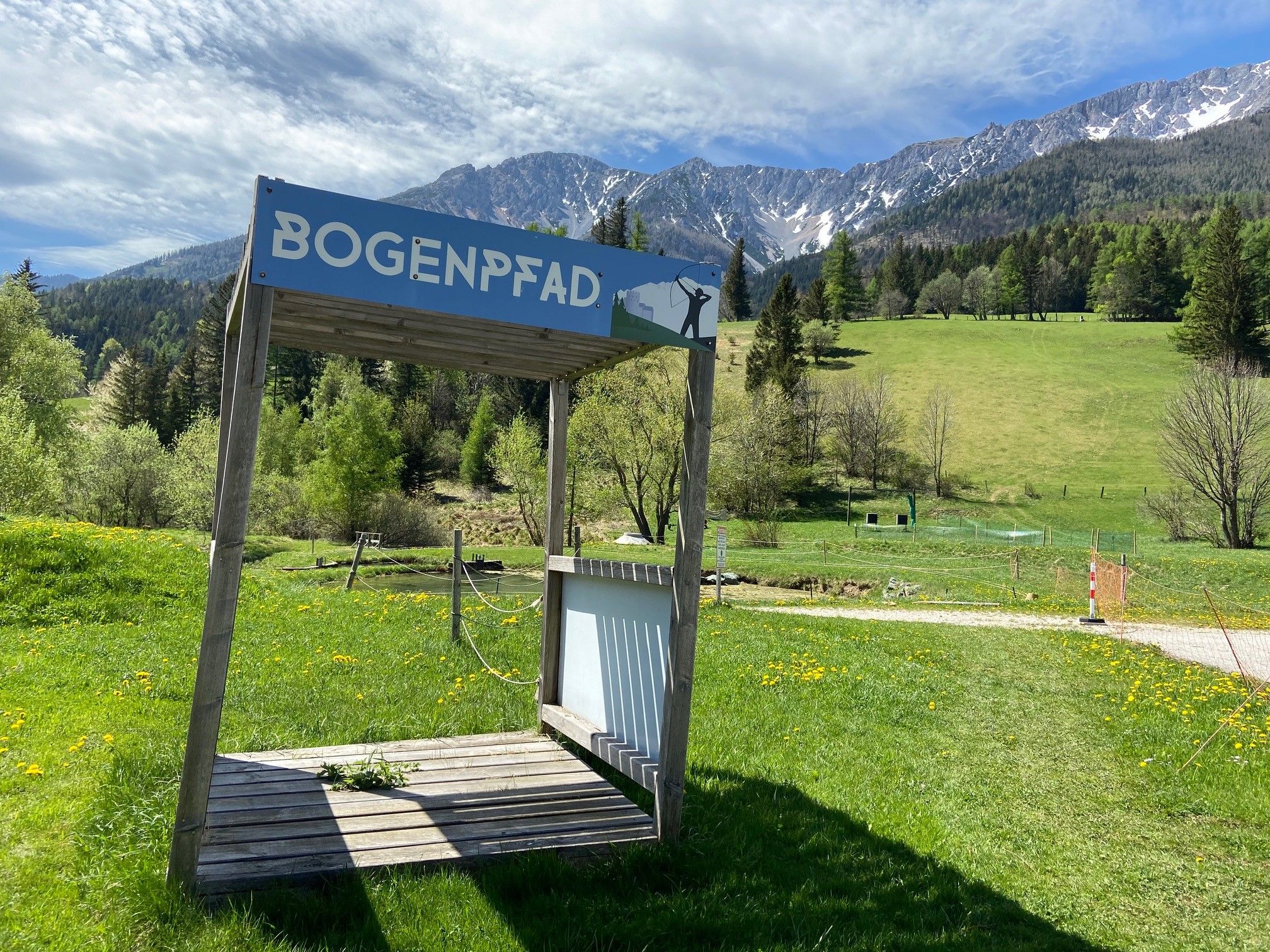 Holzplattform mit Schild 'Bogenpfad' vor einer Berglandschaft.