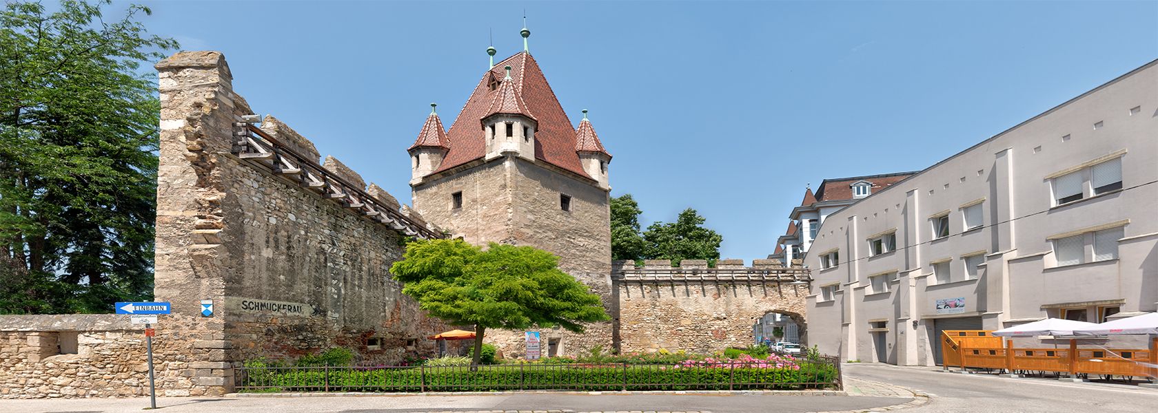Historischer Reckturm mit rotem Dach und angrenzender Stadtmauer in einer urbanen Umgebung.