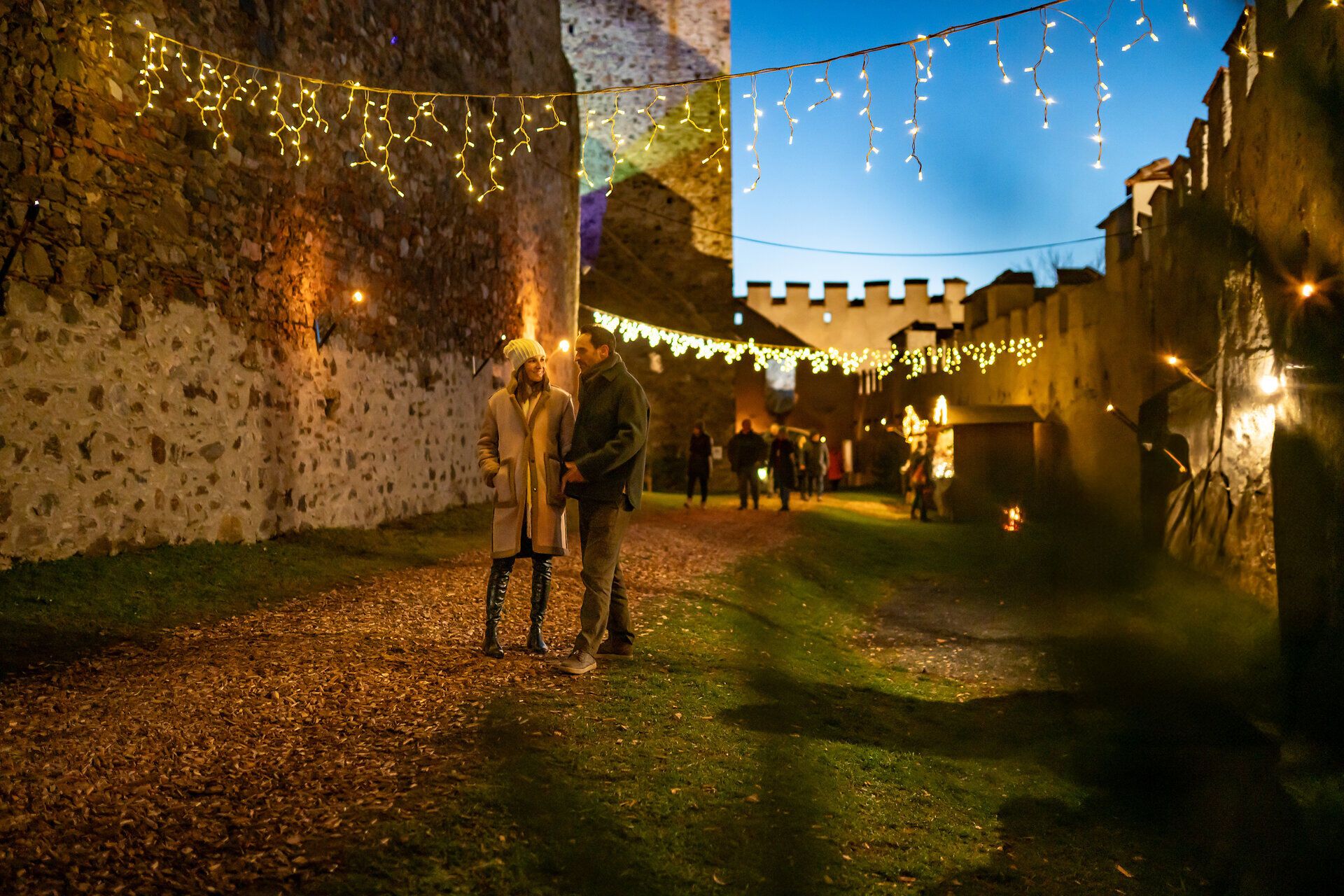 Besucher gehen durch einen festlich beleuchteten Burghof mit Lichterketten und Marktständen am Abend.