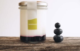 A glass filled with yogurt and fruit on a wooden table, next to it 3 stacked blueberries.