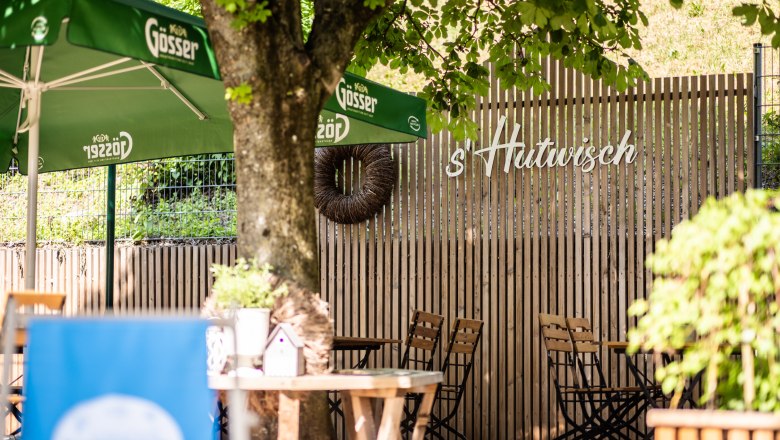 Ein Gastgarten mit Holztischen und -stühlen, grünen Sonnenschirmen und einem Holzzaun mit der Aufschrift 's'Hutwisch'. in der Mitte steht ein Baum, der von einer Bank umgeben ist.