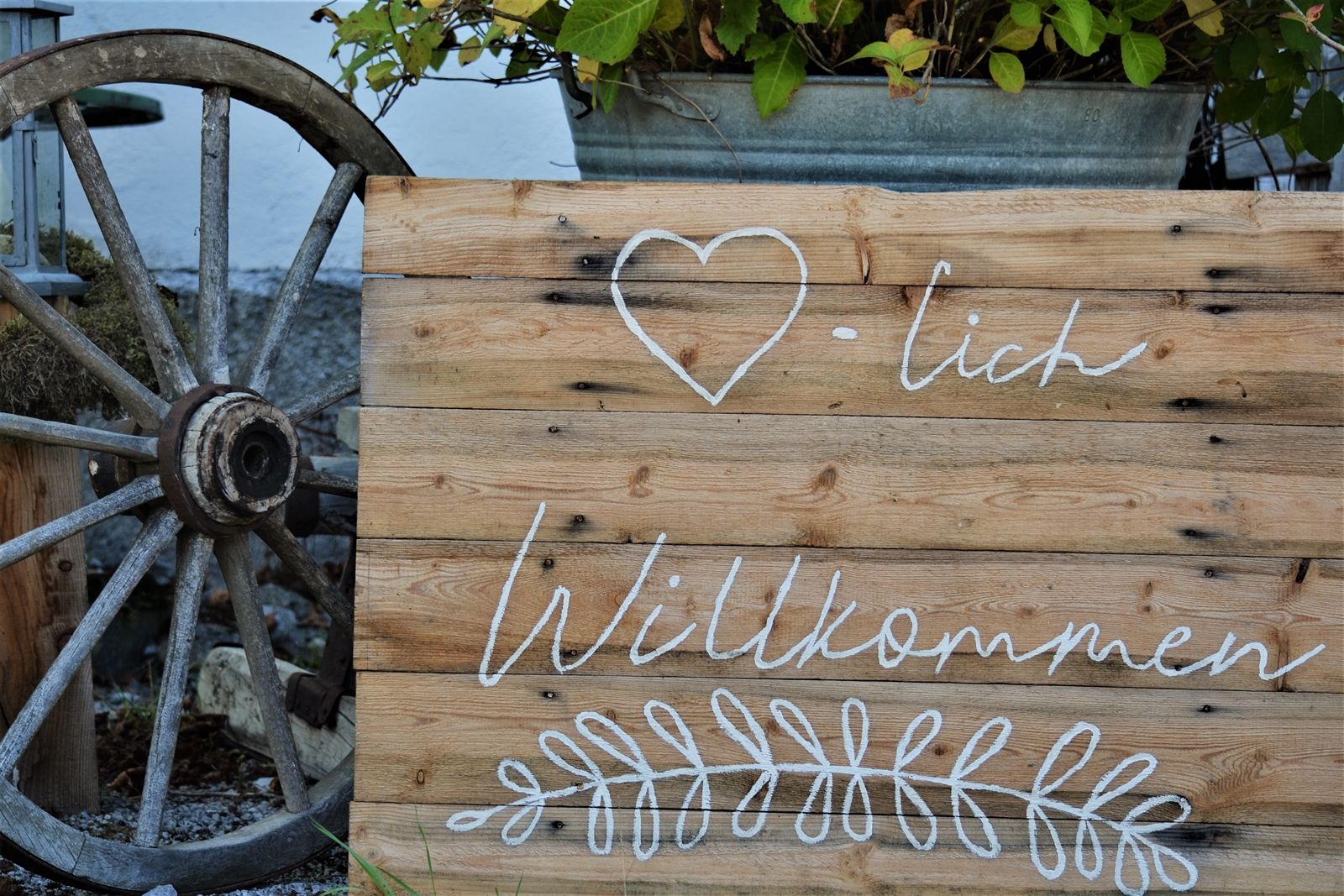 Holzschild mit der Aufschrift 'Herzlich Willkommen' und einem Herzsymbol, daneben ein altes Wagenrad.