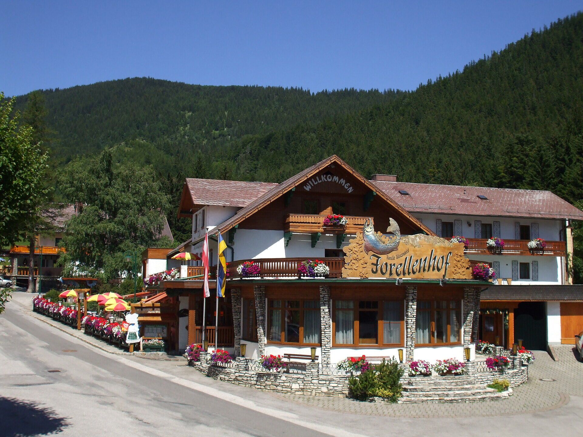 Außenansicht des Forellenhofs mit Blumenschmuck, Gastgarten und bewaldeten Berghängen im Hintergrund