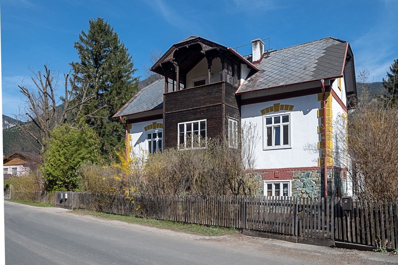Ein traditionelles Landhaus mit Holzbalkon und weißer Fassade, umgeben von Bäumen und Sträuchern, unter einem klaren blauen Himmel.
