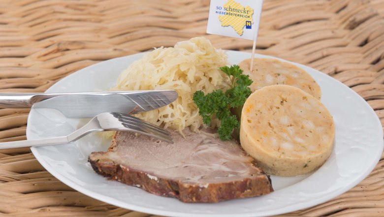 White plate on a wicker table with roast pork, dumplings and sauerkraut, garnished with parsley.