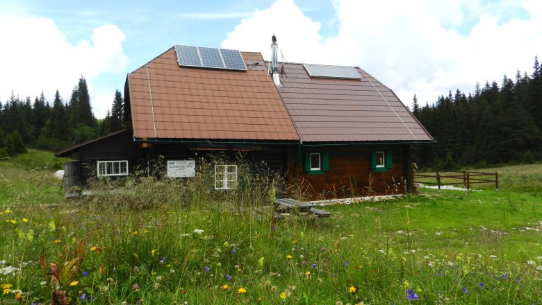 A mountain hut with solar panels on the roof, surrounded by a flowering meadow and forest in the background.