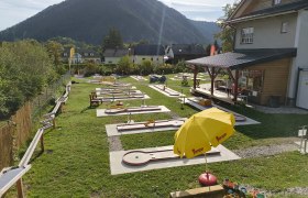 Minigolfplatz mit mehreren Bahnen und einem gelben Sonnenschirm, umgeben von Bergen und Häusern.