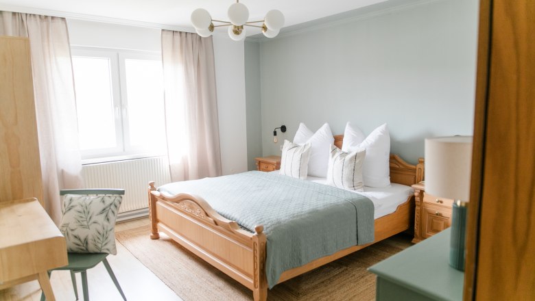 Ein gem&uuml;tliches Schlafzimmer mit einem Holzbett, blauer Bettdecke, Nachttischen und einem Schreibtisch mit Stuhl. Helle Vorh&auml;nge und ein Fenster sind zu sehen.