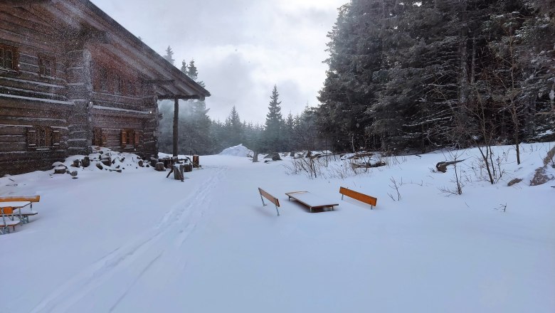 Verschneite Alm mit Holzhütte und Bänken im Winterwald.