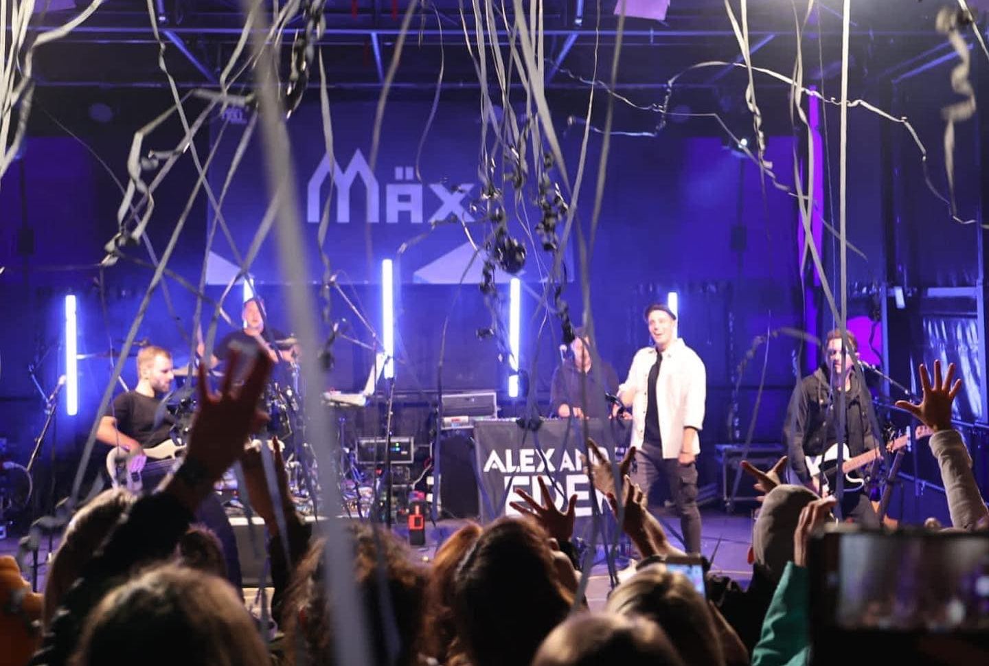 Livekonzert auf einer beleuchteten Bühne mit Band, herabfallendem Konfetti und jubelndem Publikum im Vordergrund.