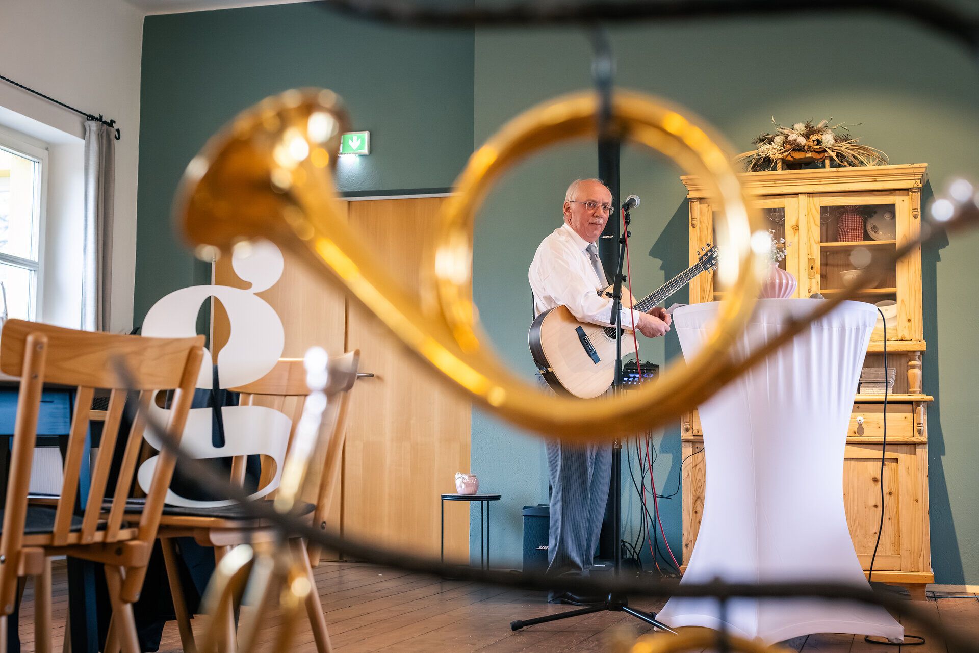Musiker mit Gitarre durch ein Blasinstrument fotografiert steht in einer Gaststube mit Blick in die Kamera