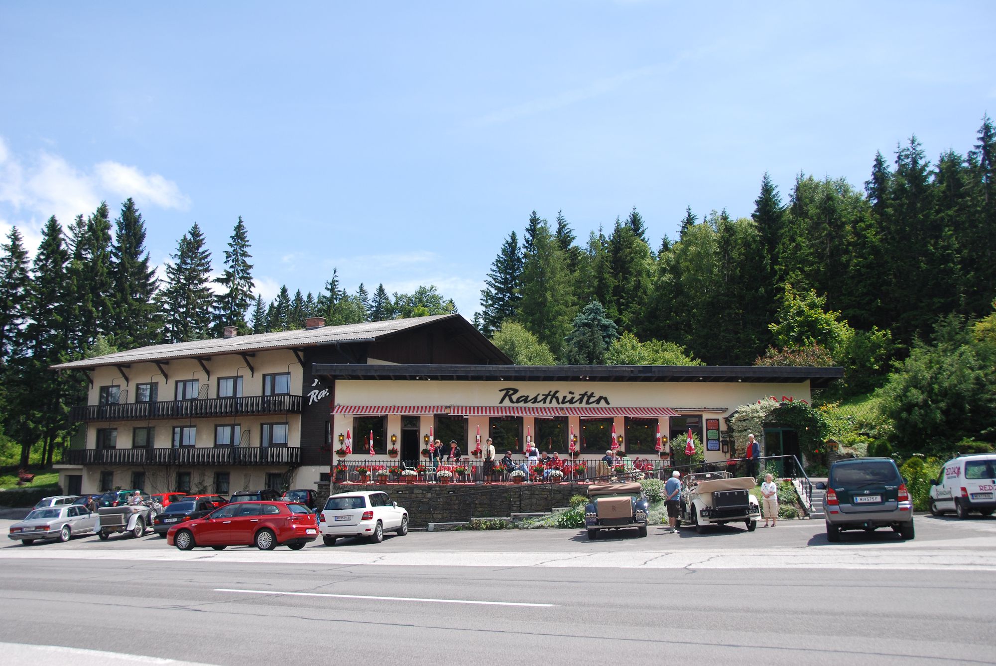 Ein Rasthaus mit Terrasse und geparkten Autos davor, umgeben von Bäumen.